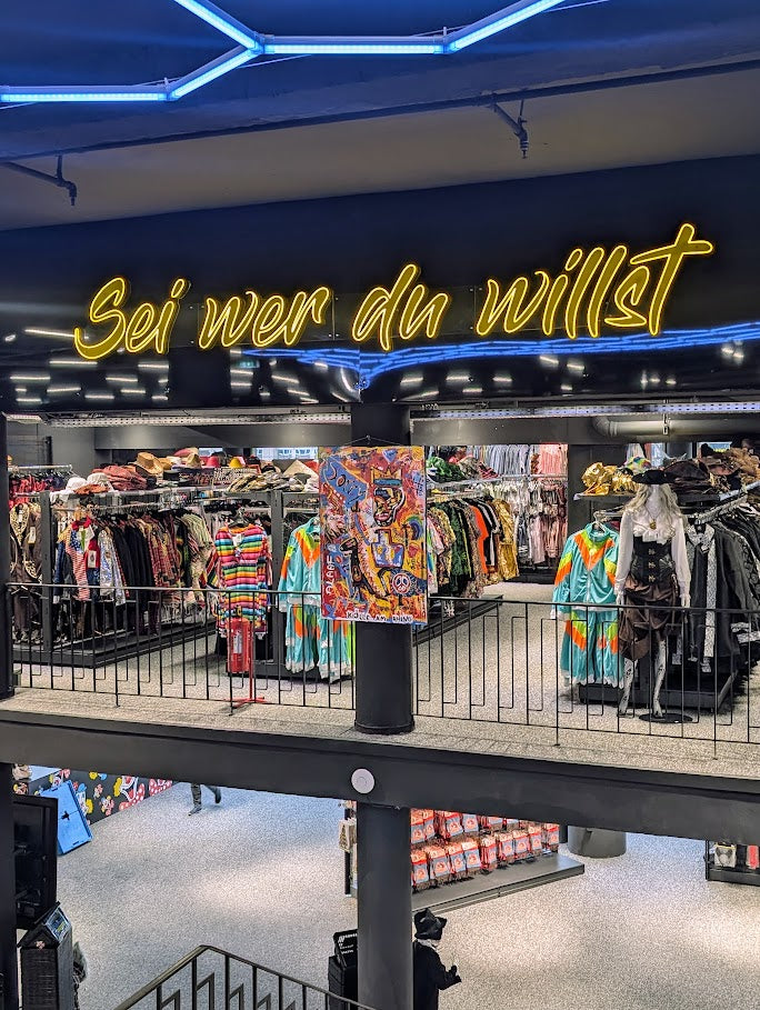 Ausstellungsansicht des Gemäldes „Die (fast) wahre Geschichte vom Aussterben der Dinosaurier“ von Jasmin Meirose, gezeigt in einem Modegeschäft mit dem Leuchtschriftzug „Sei wer du willst“. Das farbenfrohe Werk hängt zwischen bunten Kleidungsstücken und grellem Neonlicht – ein spielerischer Dialog zwischen Kunst und Popkultur.