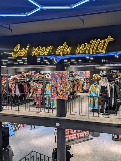 Ausstellungsansicht des Gemäldes „Die (fast) wahre Geschichte vom Aussterben der Dinosaurier“ von Jasmin Meirose, gezeigt in einem Modegeschäft mit dem Leuchtschriftzug „Sei wer du willst“. Das farbenfrohe Werk hängt zwischen bunten Kleidungsstücken und grellem Neonlicht – ein spielerischer Dialog zwischen Kunst und Popkultur.
