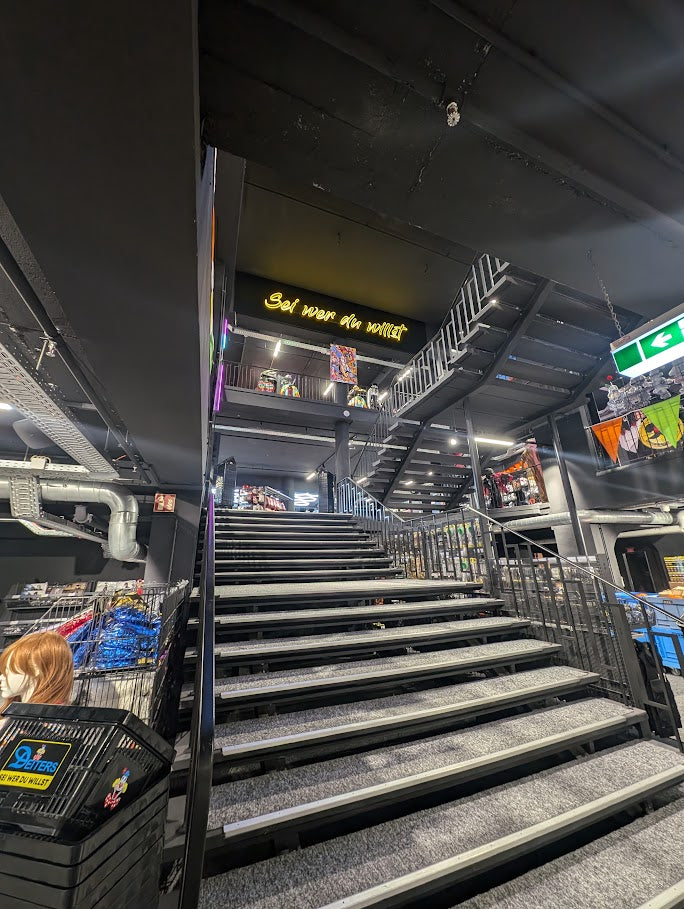 Blick auf eine Treppe in einem modernen Modegeschäft mit dem Leuchtschriftzug „Sei wer du willst“. Oben im Treppenbereich ist das farbenfrohe Gemälde „Die (fast) wahre Geschichte vom Aussterben der Dinosaurier“ von Jasmin Meirose zu sehen, als Teil einer urbanen Kunstausstellung zwischen Mode und Street-Art-Ästhetik.