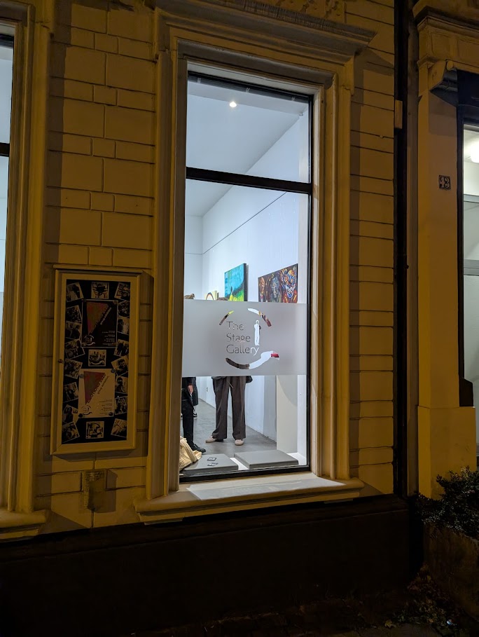 Außenansicht einer Galerie bei Nacht mit Blick durch das Fenster auf die Ausstellung „Dinner for (every)one – ein Anti-Abendmahl“ von Jasmin Meirose. Das Triptychon ist im Innenraum beleuchtet sichtbar, Besucher betrachten das Werk.