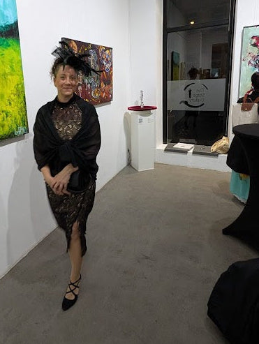 Galerieansicht während der Ausstellung von Jasmin Meirose. Im Vordergrund steht die Künstlerin elegant gekleidet im Raum, im Hintergrund ist ihr Werk „Dinner for (every)one – ein Anti-Abendmahl“ an der Wand sichtbar.