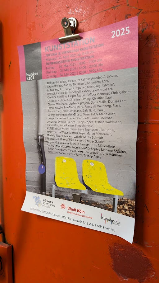 Plakat der Kunstroute Ehrenfeld 2025, befestigt an einer roten Wand mit Metallclip. Zu sehen sind die Logos der Stadt Köln, der Käthe-Kollwitz-Stiftung und der Kunstroute Ehrenfeld. Unter den teilnehmenden Künstlerinnen und Künstlern ist auch Jasmin Meirose aufgeführt.